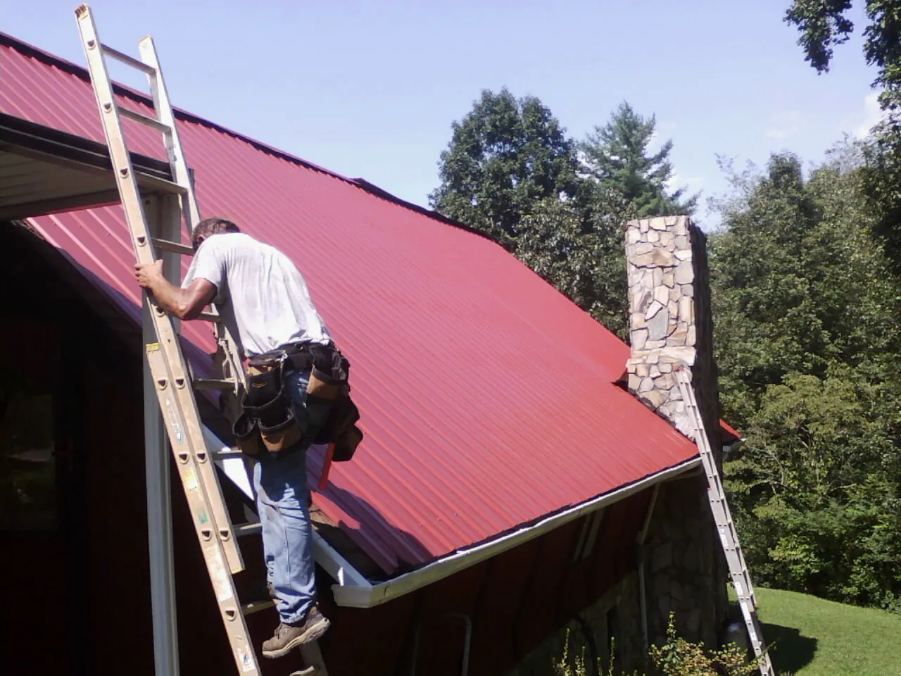 Roofer on a ladder beside a red metal roof and stone chimney — Soffit & Fascia Repair by Roofing Brothers in Natrona Heights